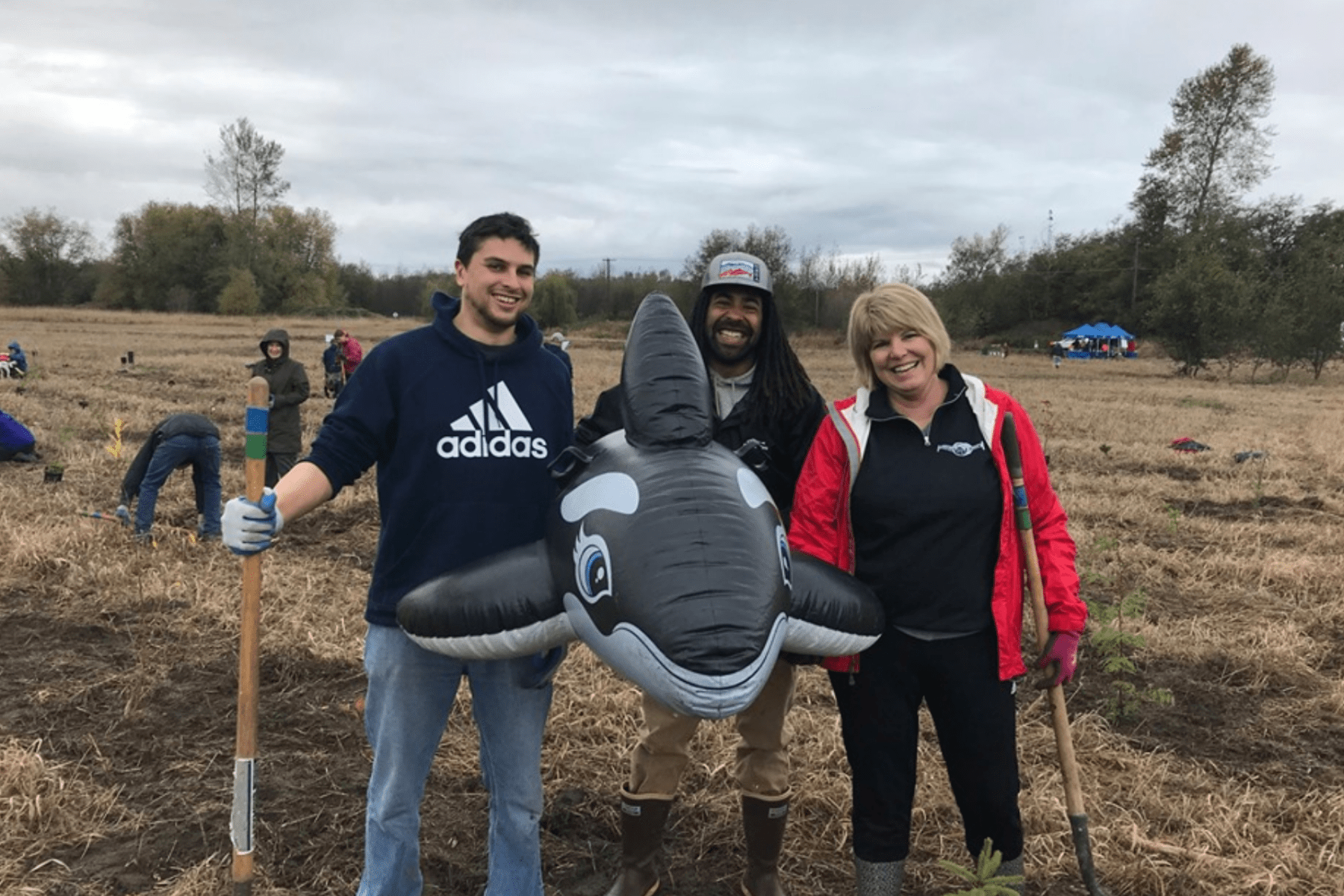 People holding shovels and a blow up orca whale while standing in a field 