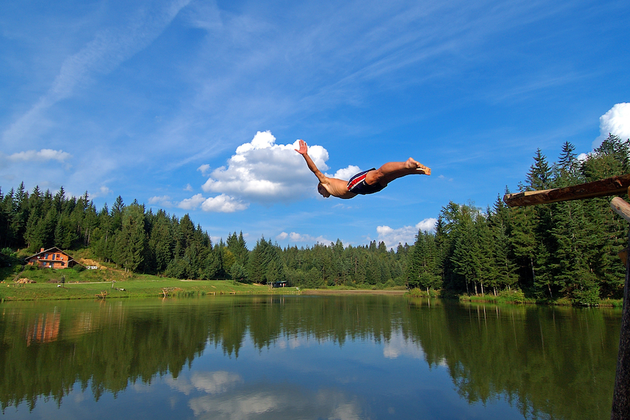 Man in blue shorts leaping belly first into a lake surrounded by green trees 