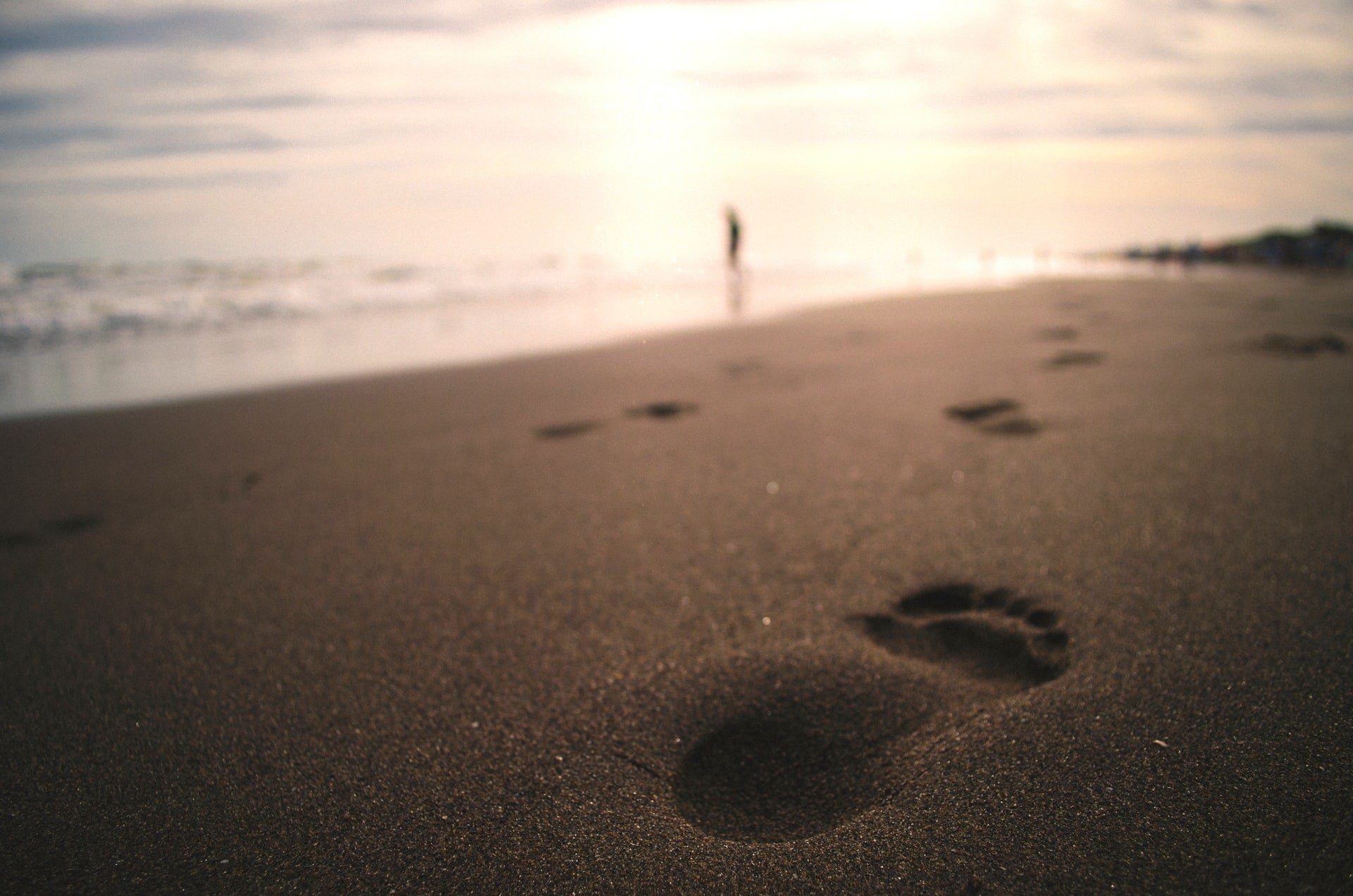 dark foreground medium brown sand with dark footprints ocean and setting sun in distance blurred human figure far away