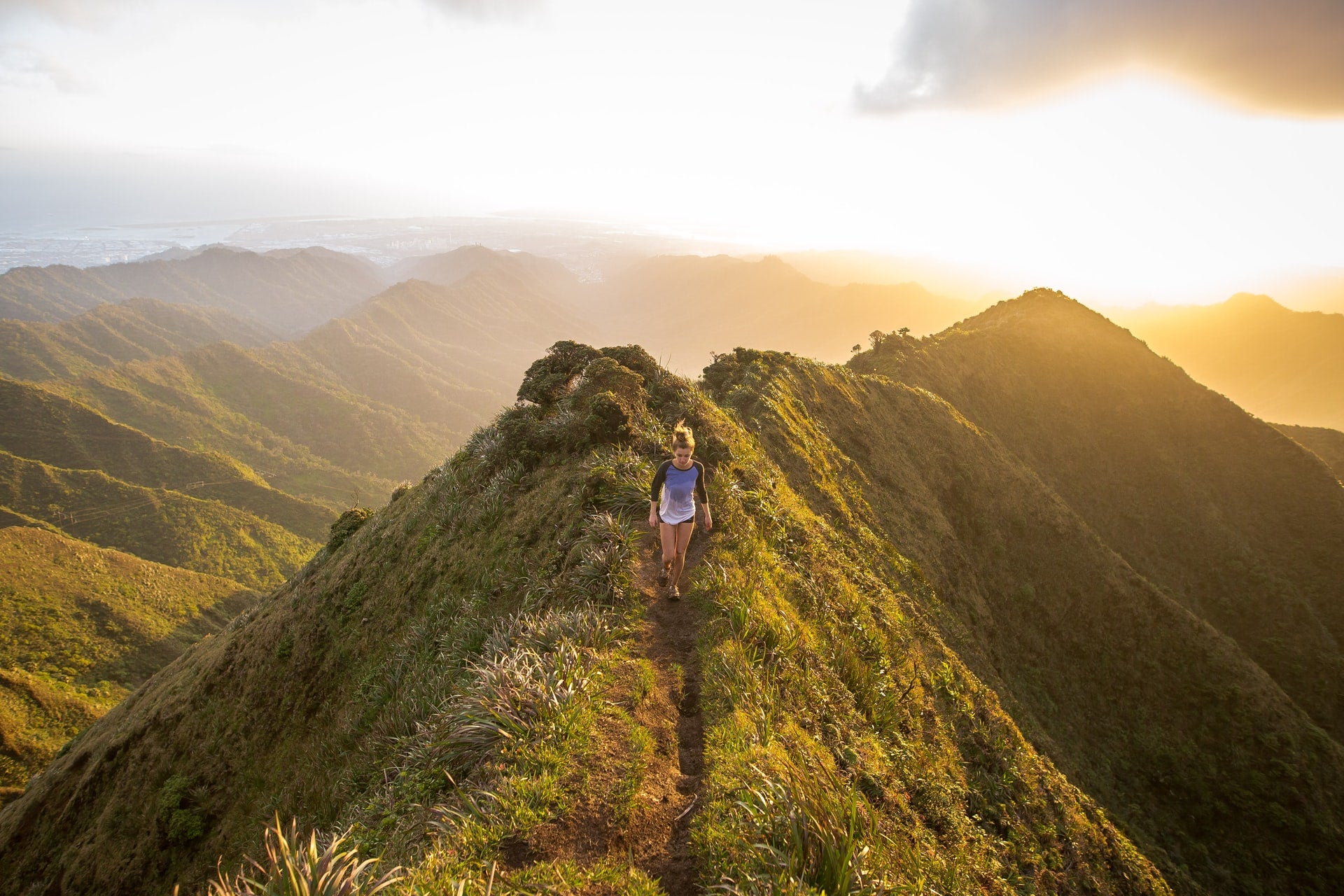 young woman hiking on lush green mountaintop with sunrise and distant mountain ranges in background