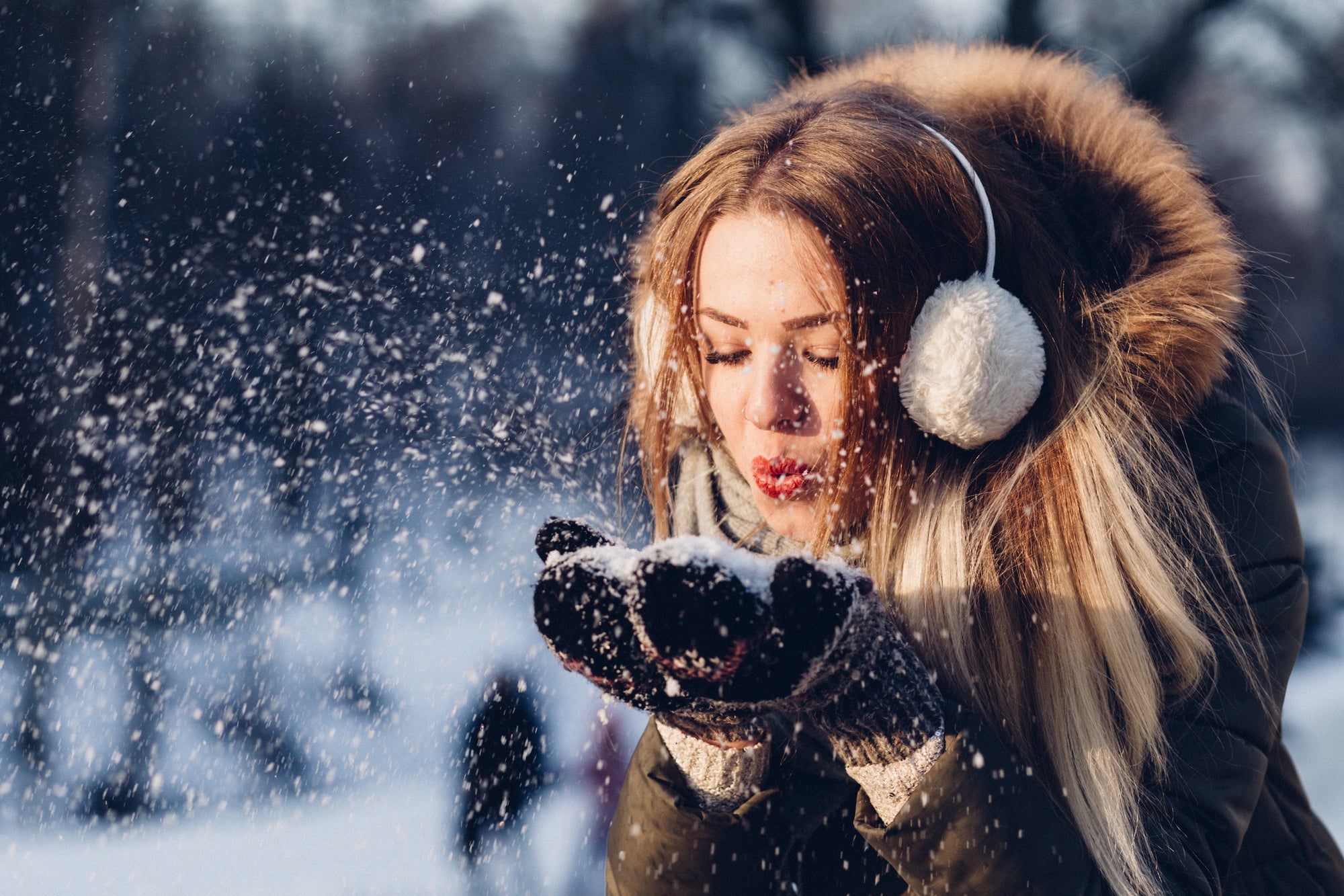 woman with white ear muffs and furry winter jacket blows white snow from her mittens 