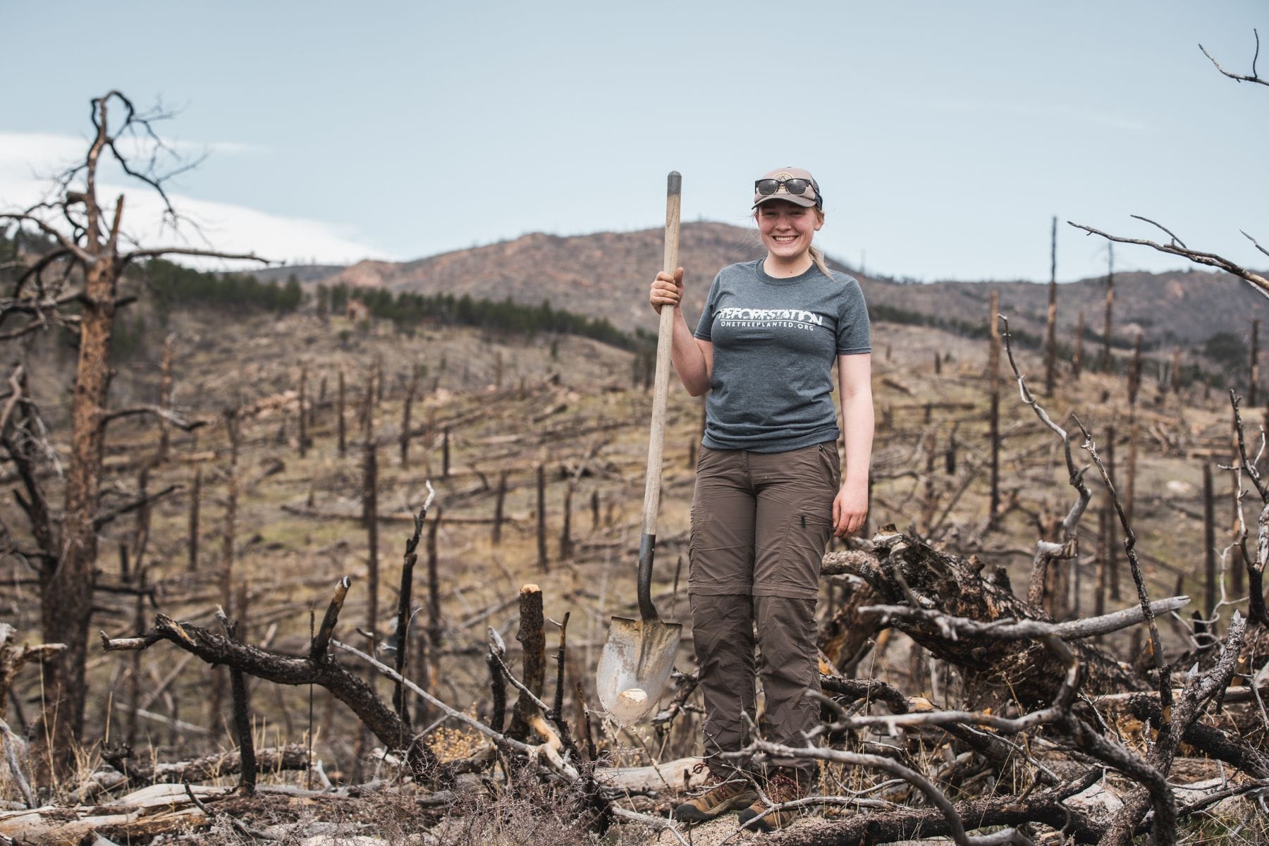 woman holding shovel, smiling at a site where she is planting trees for wildfire restoration in colorado. surrounded by burned trees